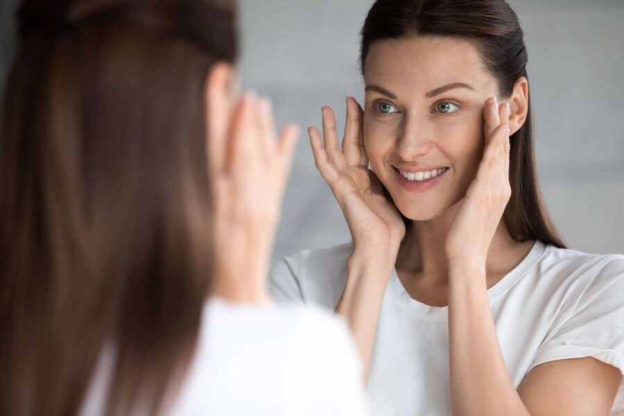 Smiling young brunette woman looking at mirror, touching face.