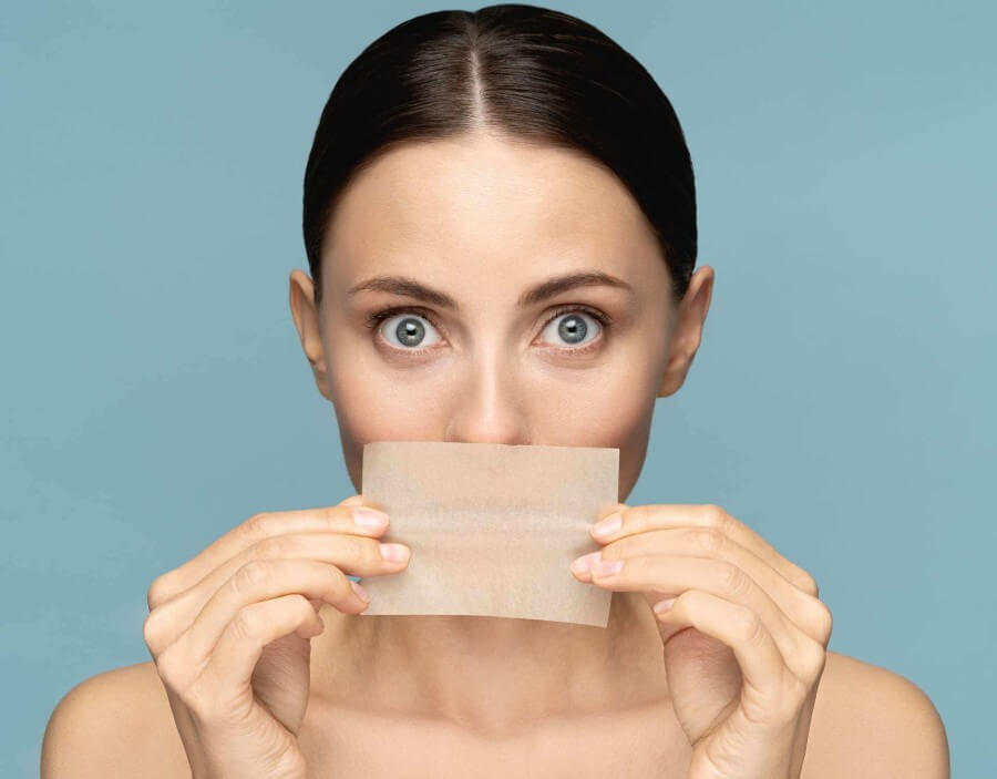 close-up-of-woman-with-natural-face-makeup-holding-facial-oil-blotting-paper-hides-her-lips-looking_t20_3gyj99