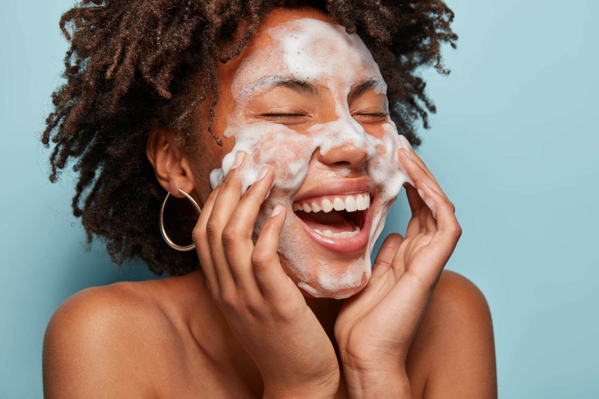 cheerful black female model applies foaming cleanser, has clean fresh healthy skin, smiles broadly, afro bushy haircut, stands bare shoulder against blue background. beauty and feminine concept