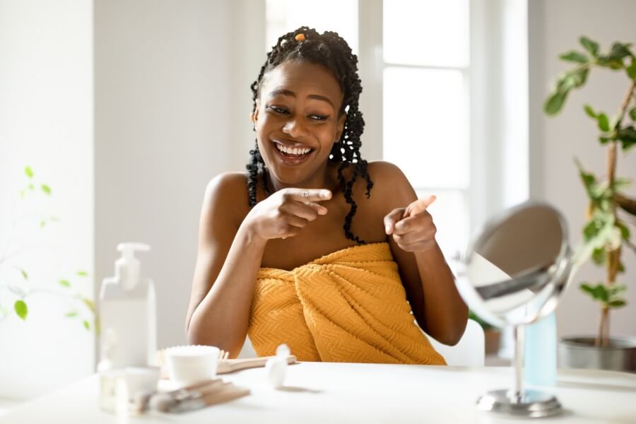 power of daily skincare. happy african american lady pointing fingers at her reflection in mirror, sitting in bedroom