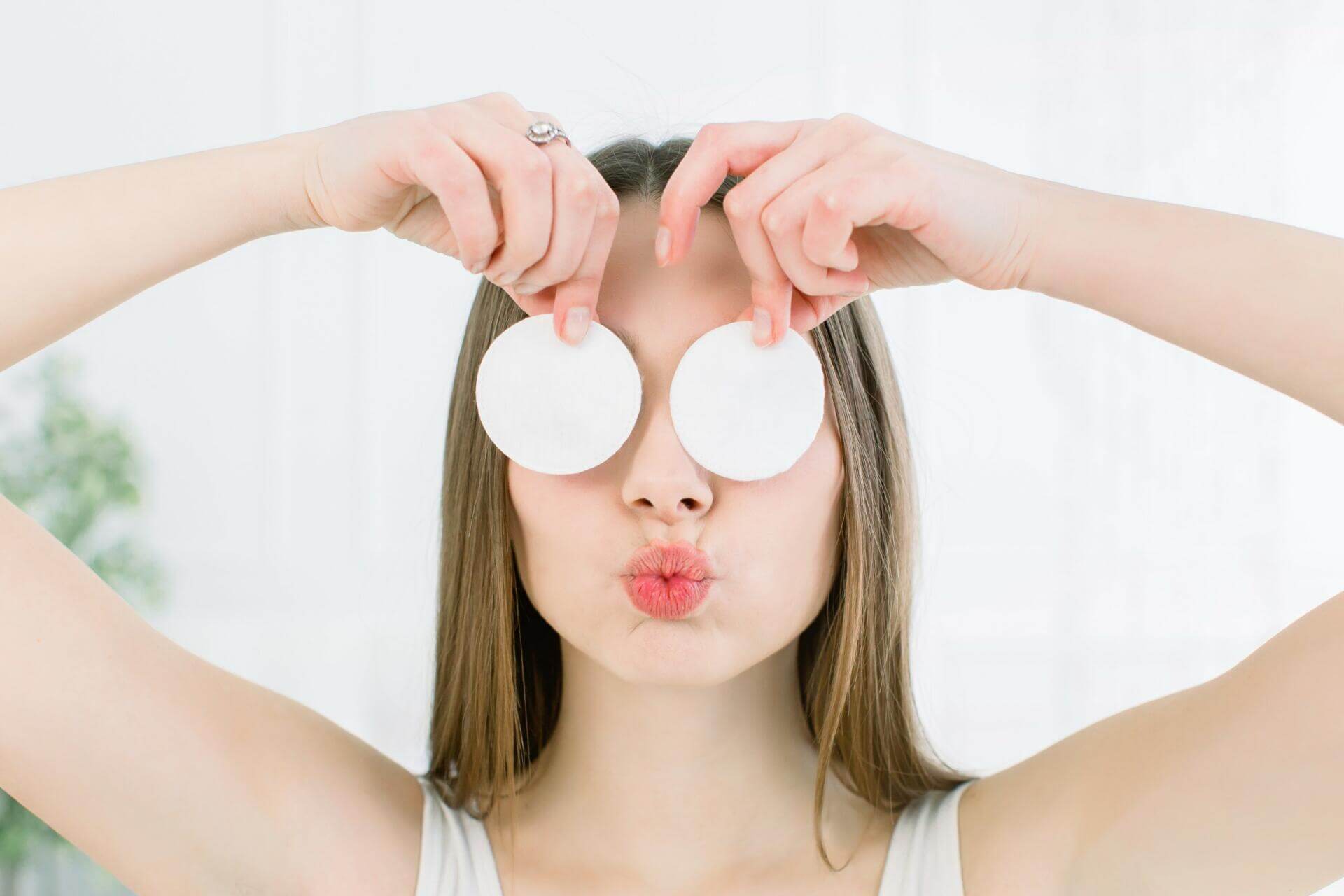 happy and positive young beautiful woman with open shoulders covering her eyes with a cotton pads and showing kiss lips on a light background. skin care and beauty concept