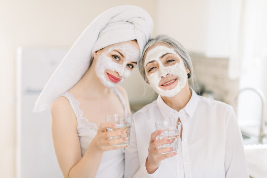 beauty, skin care and healthy lifestyle concept. indoor home shot of two women, mother and daughter, relaxing with facial masks, posing to camera with glasses of water over light background