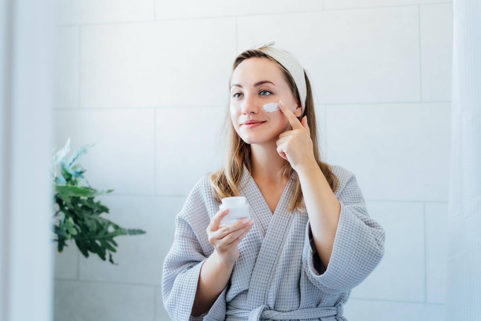 young woman in bathrobe looking in the mirror, holding bottle with moisturizer cream and applying it on face. skin hydrating. cosmetic spa procedures. beauty self care at home. selective focus.