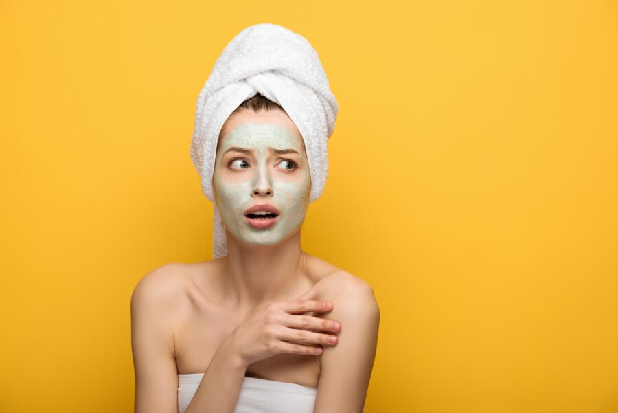 scared girl with nourishing facial mask and towel on head looking away isolated on yellow