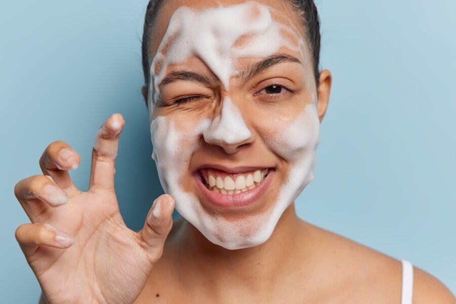 close up shot of positive woman undergoes facial skin care routine keeps hand as if palm winking eye shows white teeth isolated on blue background. daily hygiene treatments and complexion care concept