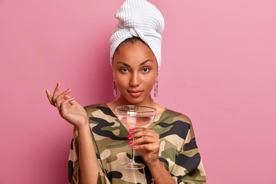 horizontal shot of serious dark skinned female model looks confidently at camera raises hand dressed in domestic outfit with wrapped towel on head drinks cocktail poses against pink background