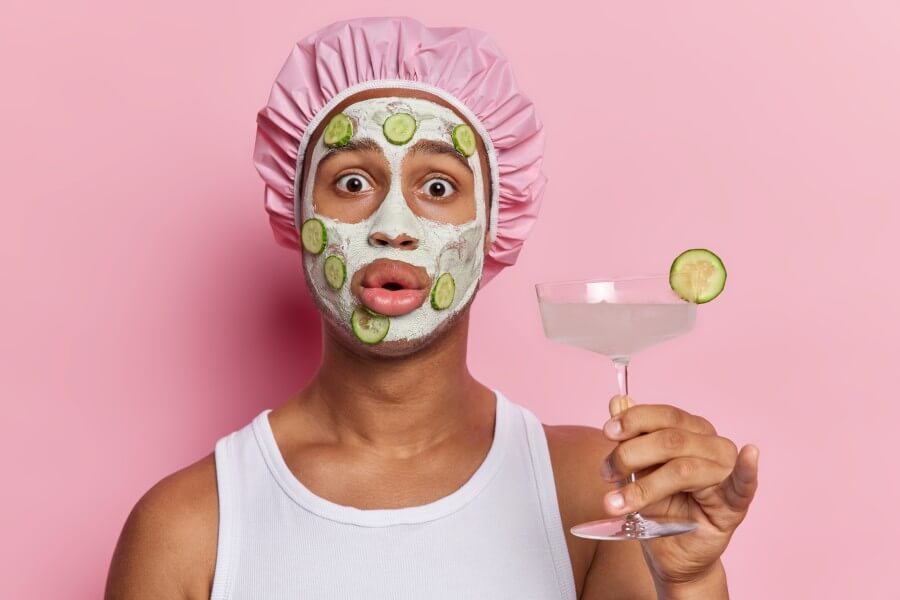 impressed adult man holding refreshing cocktail gently applies facial clay mask accompanied by soothing cucumber slices stares startled wears white t shirt and bath hat isolated over pink background