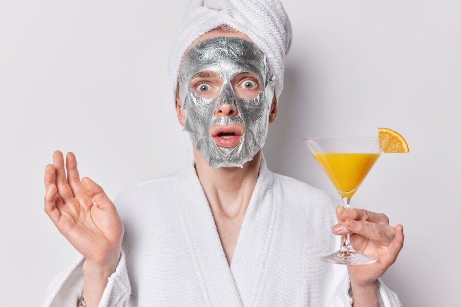 scared surprised man applies beauty silver mask stares impressed at camera wears bath towel on head and robe holds orange cocktail undergoes beauty procedures for skin care isolated over white wall