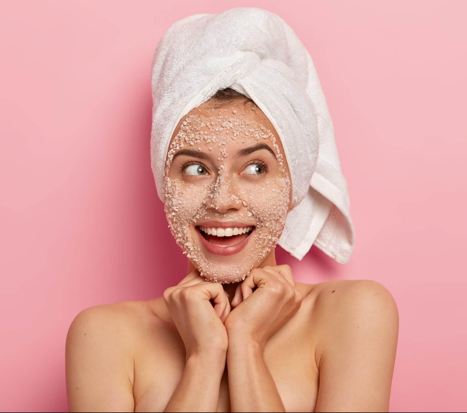 beauty and feminity concept. pretty cheerful female keeps both hands under chin, has dreamy thoughtful expression, wears white soft towel on head, has naked body, applies organic sea salt scrub