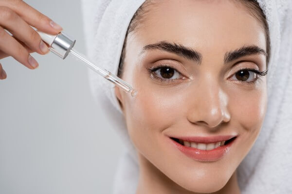 close up of happy young woman applying moisturizing serum with pipette isolated on grey