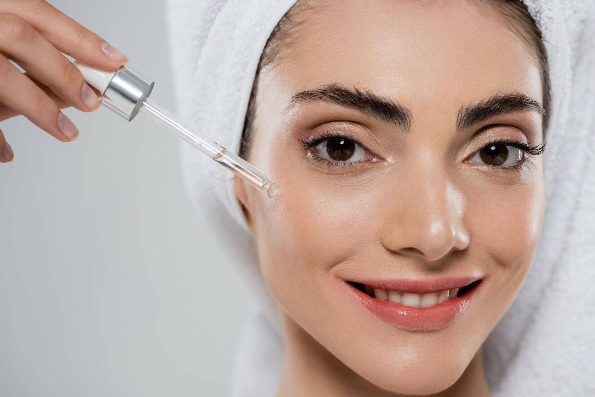 close up of happy young woman applying moisturizing serum with pipette isolated on grey