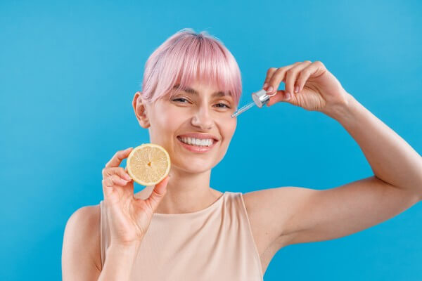 portrait of smiling woman with pink hair holding half of fresh lemon and a dropper near her face isolated over blue background. vitamin c serum