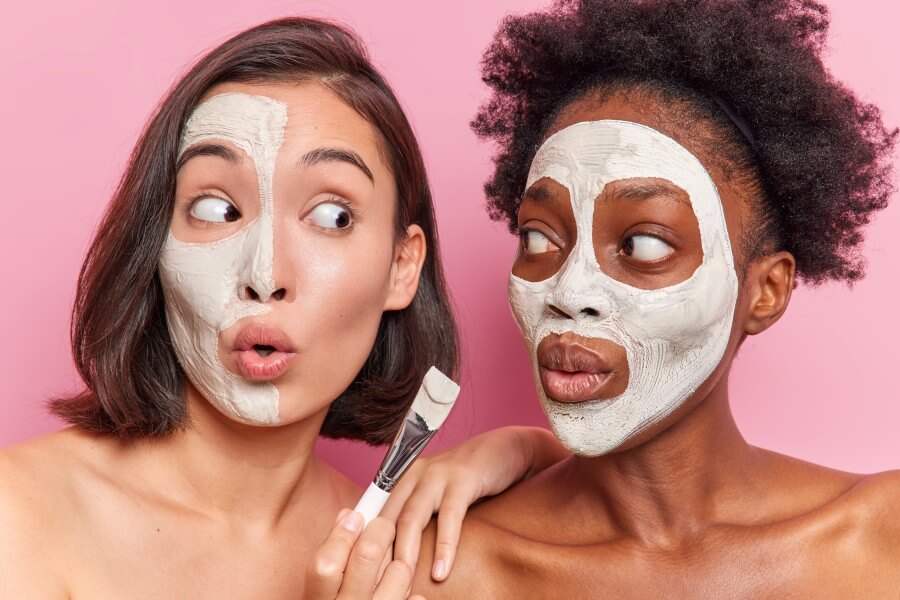 surprised mixed race friendly women apply nourishing face masks with cosmetic brush look shocked at each other stand shirtless against pink background. skin care beauty and wellness concept.