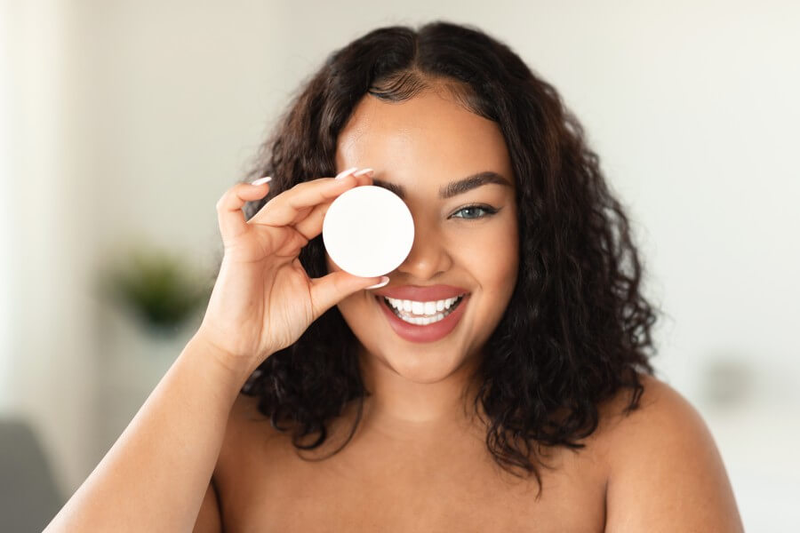 beauty products. excited black bodypositive lady holding jar with nourishing cream, covering one eye with skincare item