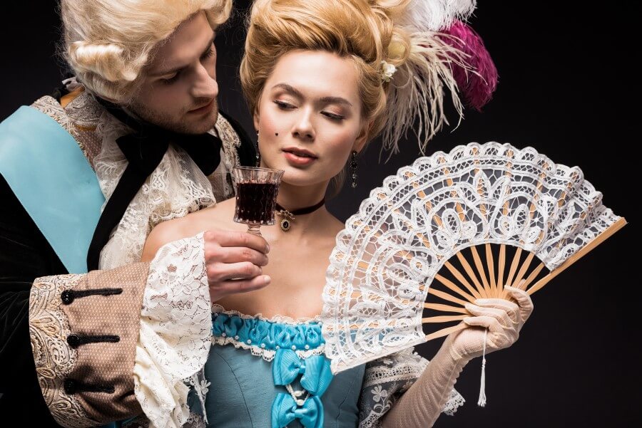 handsome man looking at young victorian woman in wig holding fan and wine glass isolated on black