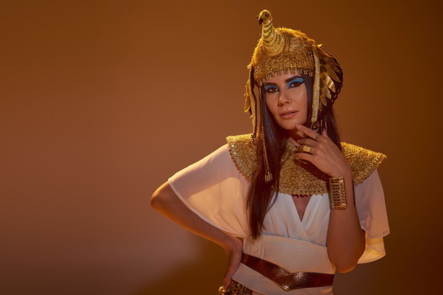 stylish woman in egyptian attire and headdress touching chin and posing on brown background