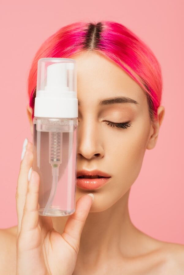 young woman with colorful hair and closed eyes holding bottle with cleansing product isolated on