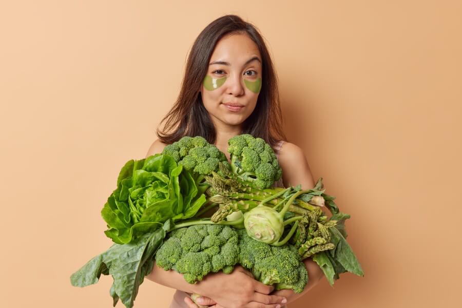 asian woman with dark hair looks seriously at camera raises eyebrows carries big heap of fresh vegetables applies hydrogel patches under eyes to reduce wrinkles poses against beige background