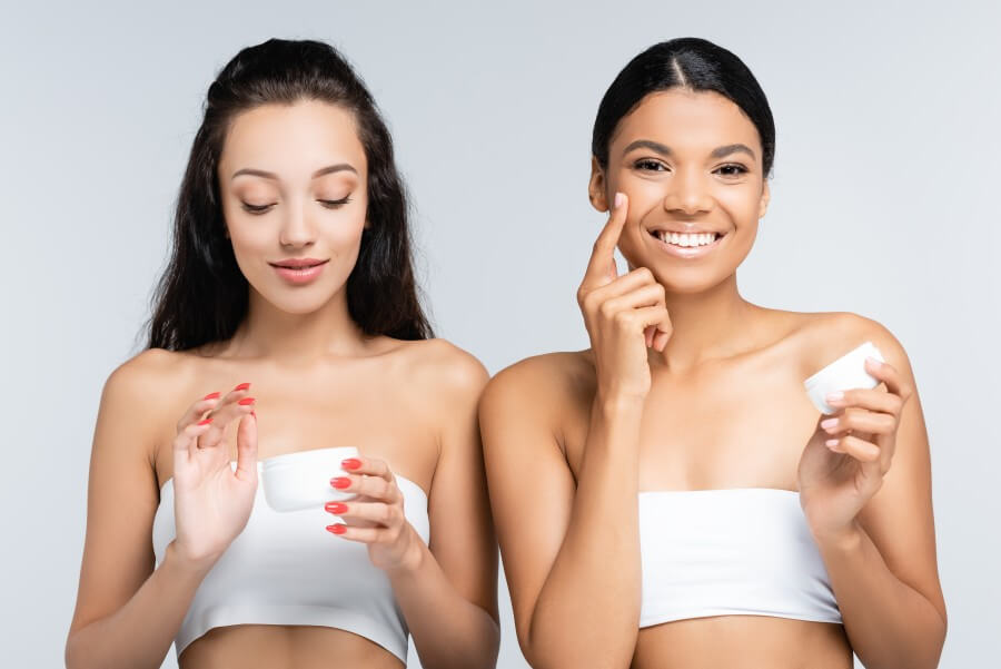 brunette multiethnic women with bare shoulders smiling while holding containers and applying