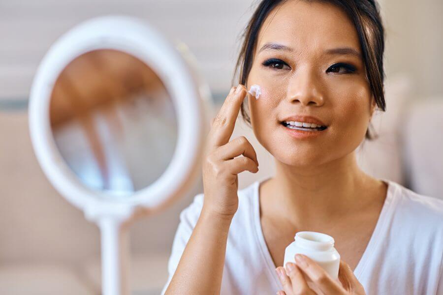 beautiful asian woman applying face cream while looking herself in a mirror.