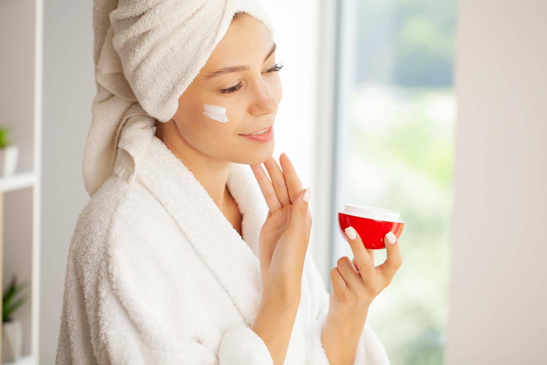 joyful young woman applying moisturizing cream on face