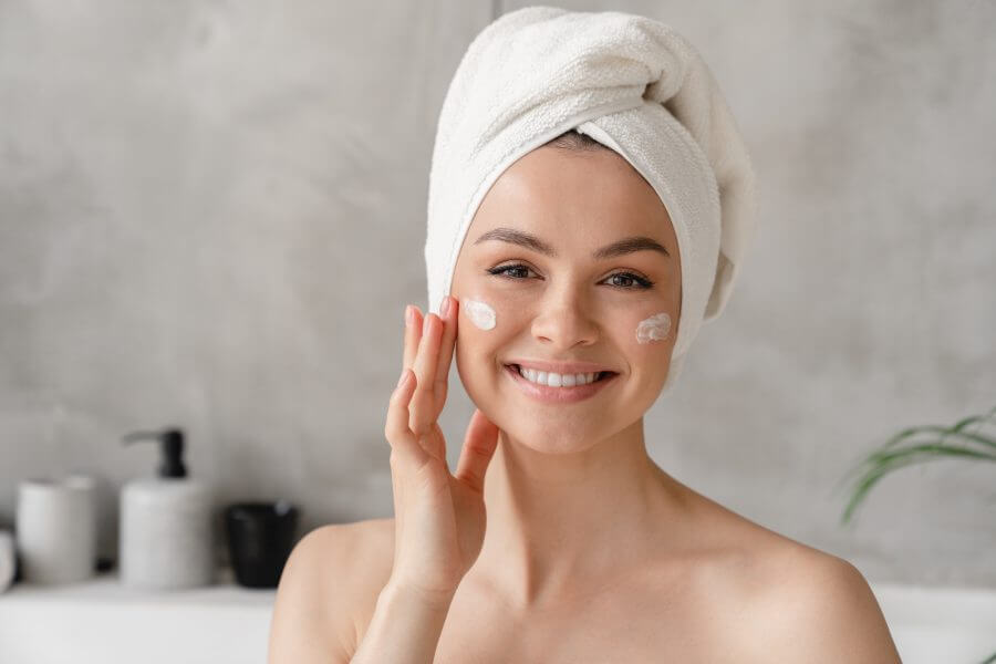 young beautiful woman in bath towel smiling applying face body cream on her face