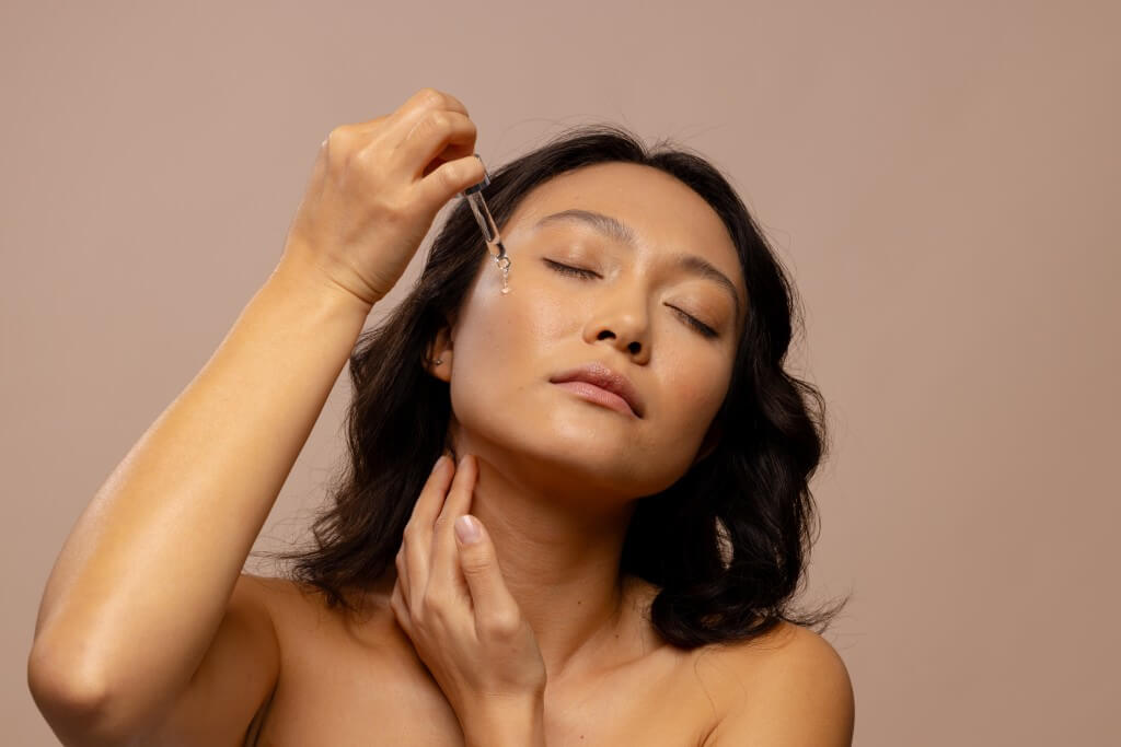 asian woman with closed eyes applying skin tonic to her cheek with dropper cap. femininity, body, skin and beauty treatments, unaltered.