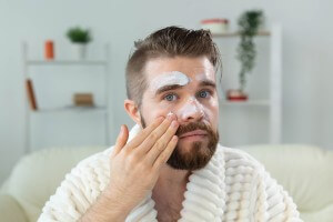 bearded guy applying facial cream in front of mirror skin care and spa for man concept.