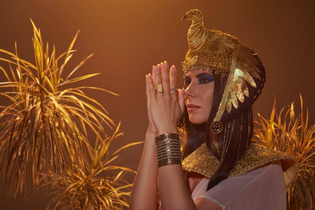 brunette woman in egyptian look doing praying hands gesture near desert plants isolated on brown