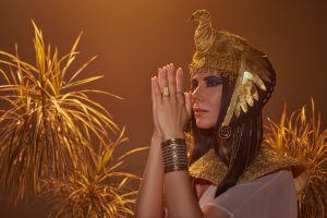 brunette woman in egyptian look doing praying hands gesture near desert plants isolated on brown