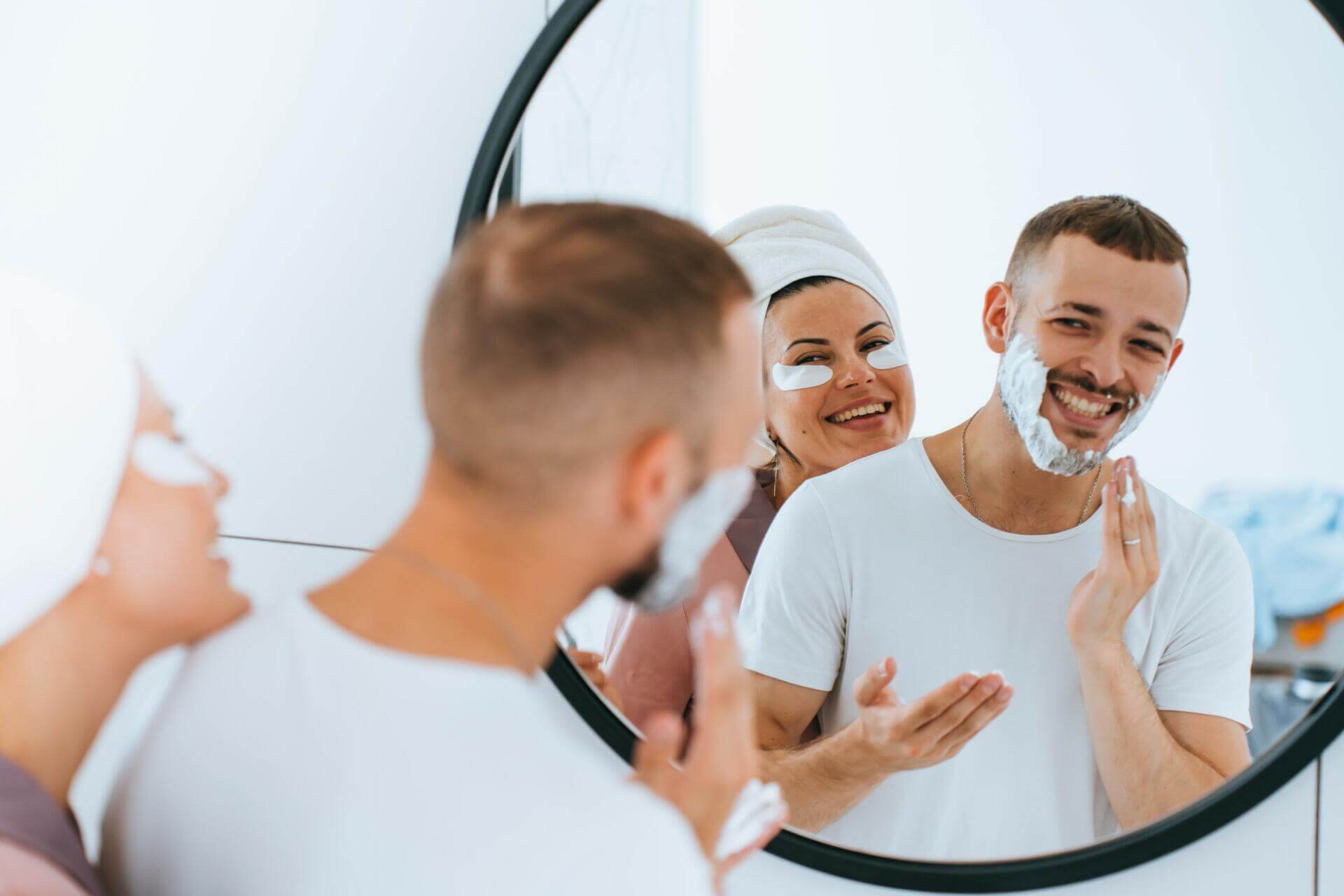 shaving beardy caucasian guy stands at mirror smiles looks at wife with cosmetic patches on face