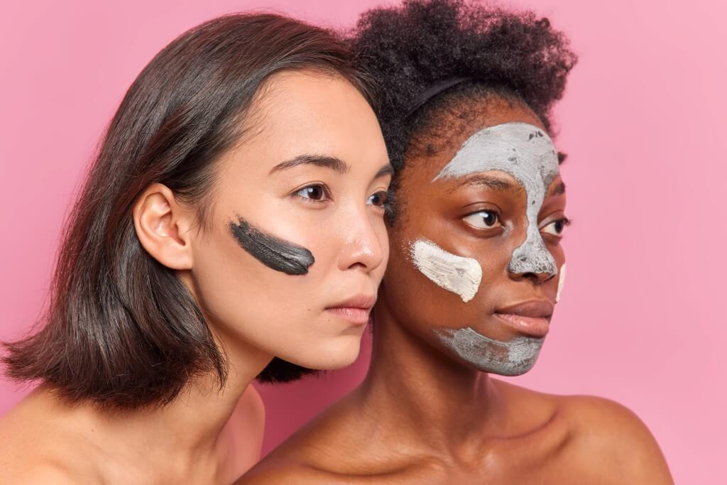 sideways shot of serious multiethnic women concentrated into distance apply clay mask on faces undergo beauty procedures prepare for date want to look young stand shirtless against pink wall