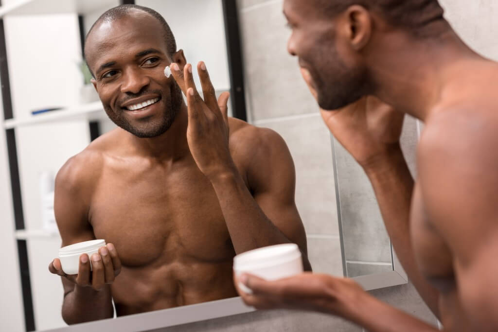 smiling young man applying facial cream while looking at mirror in bathroom