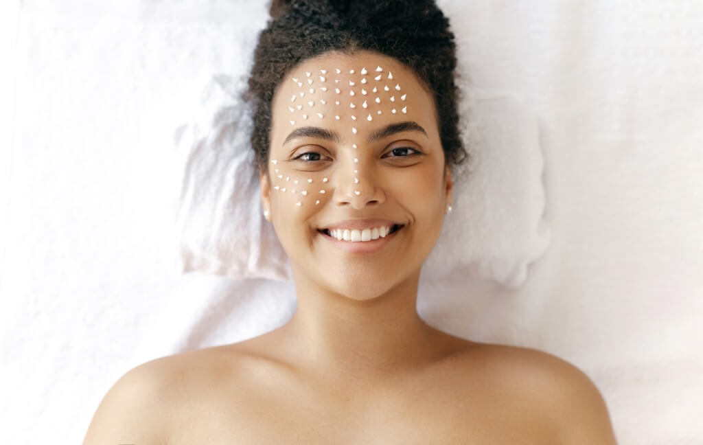 top view of a beautiful smiling hispanic or brazilian woman, with naked shoulders, lying during a skincare procedure with a cosmetologist, on her face the drops of moisturizing cream. skincare concept