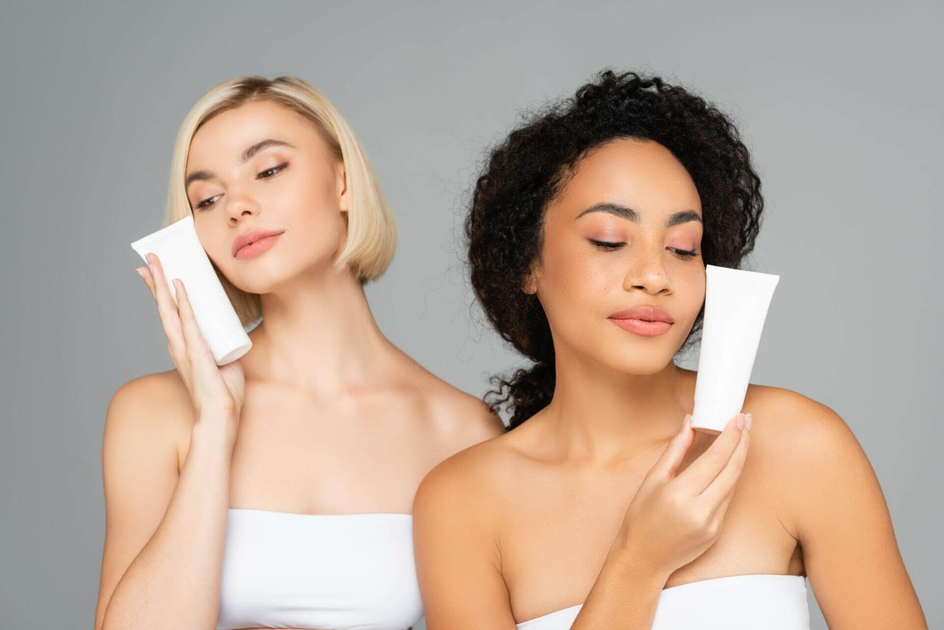 young multiethnic women holding tubes with cleansing foam isolated on grey